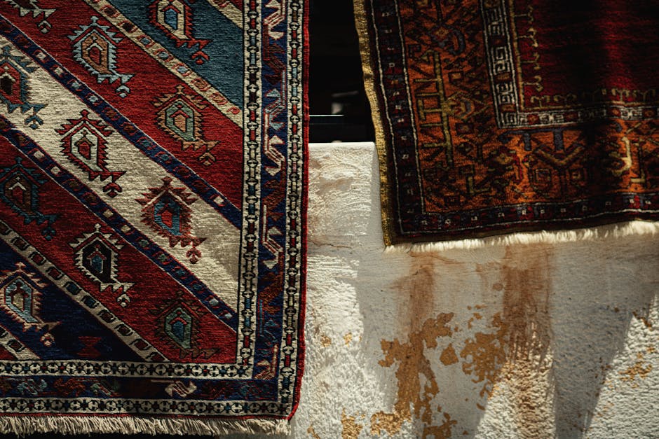 A variety of intricately patterned Persian and Oriental rugs and carpets draped over a sofa, with some rugs hanging down and others folded. The rugs feature vibrant colors including red, blue, cream,